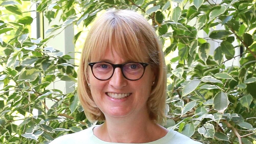 Foto einer lächelnden Frau mittleren Alters mit blondem, schulterlangem Haar, schwarz gerahmter Brille und Steckohrringen.